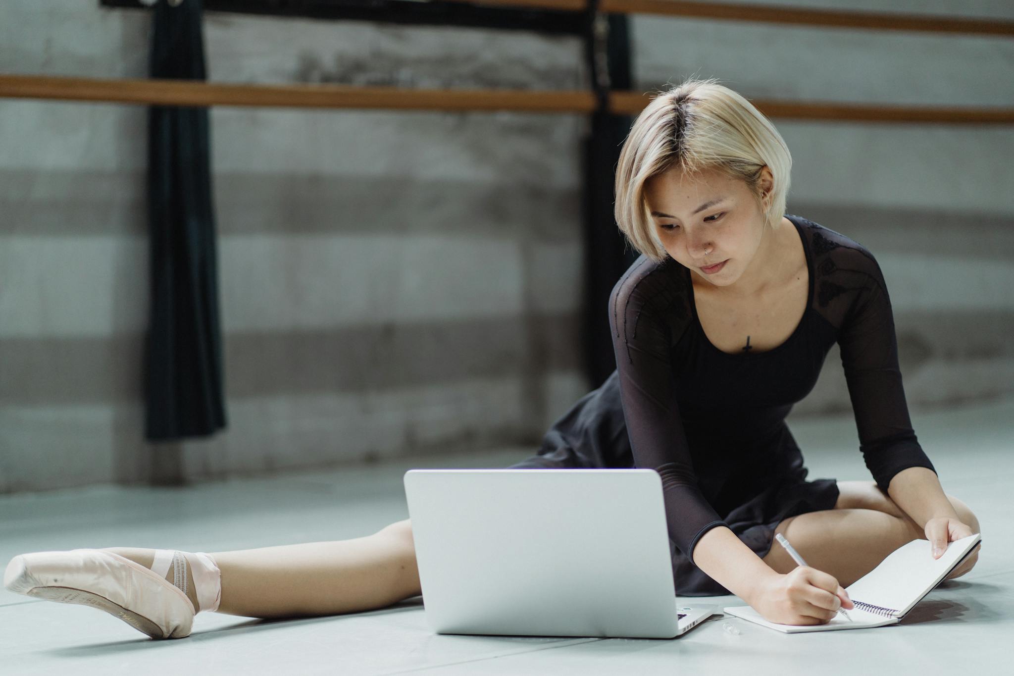 Asian ballerina studying on a laptop while taking notes in a dance studio.