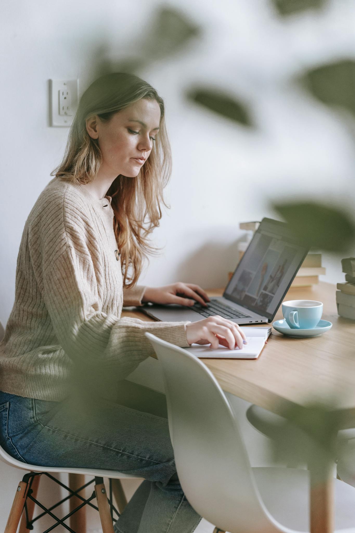 Side view of focused female freelancer reading notes in notepad while browsing netbook at table in room with blurred green leaves